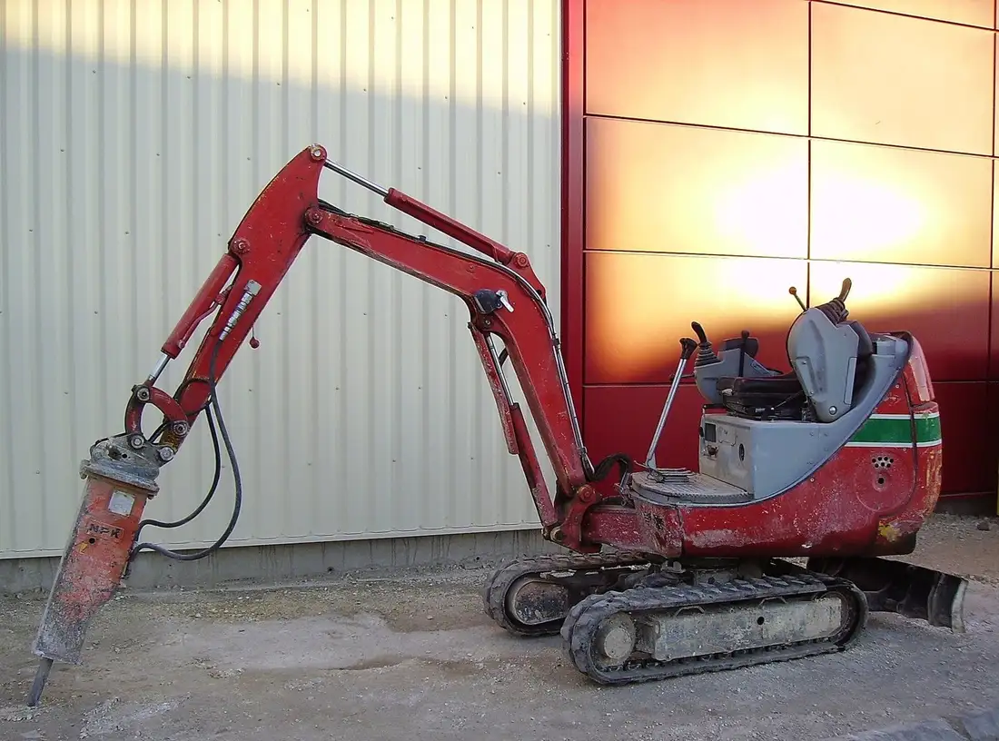 Mini-pelle rouge sans cabine, siège vide, avec un brise-roche hydraulique au bout du bras, posée sur un sol gravillonné devant un bardage