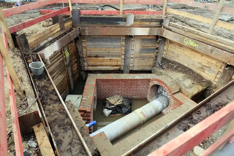 Grosse canalisation en béton raccordée à un regard en briques au fond d'une fouille blindée par des planches et des étais