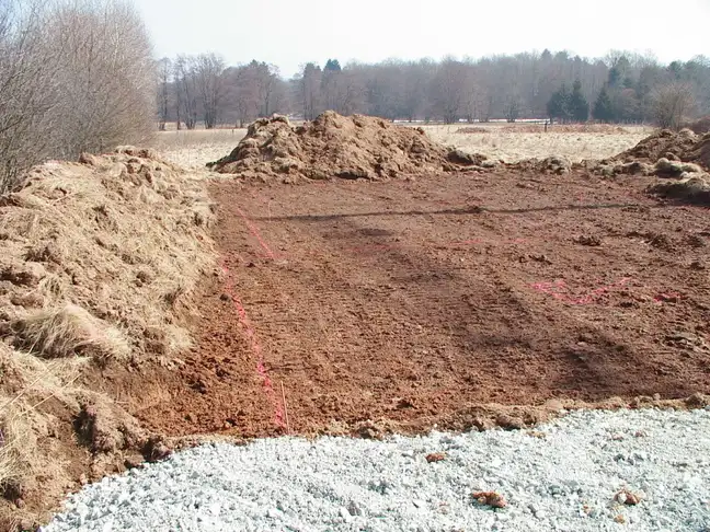 Terrain décapé en terre rouge avec tracés de piquetage rose, tas de terre herbeuse sur les côtés, gravier au premier plan