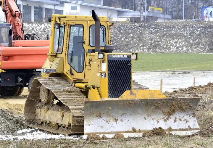 Bulldozer à chenilles garé sur un terrain en préparation, lame couverte de terre, pelle et tas de graviers derrière