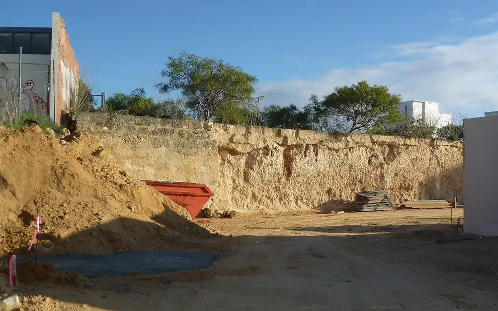 Paroi de calcaire mis à nu en bord de chantier, tas de terre ocre et benne rouge au pied