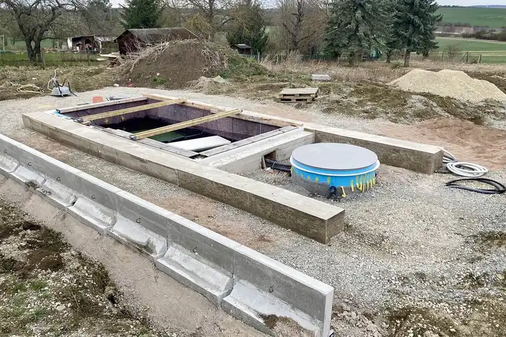 Bassin de piscine en cours de construction dans une fouille au milieu d'un terrain, coffrage bois, local technique rond bleu à côté et gravier autour