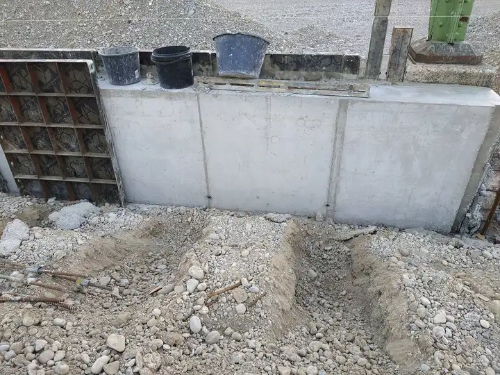Muret de béton banché avec seaux de maçon posés dessus, tranchées étroites creusées dans le gravier au premier plan