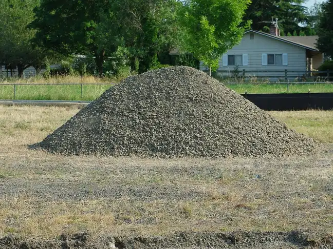 Tas de gravier gris concassé sur un terrain herbeux, maison et arbres en arrière-plan