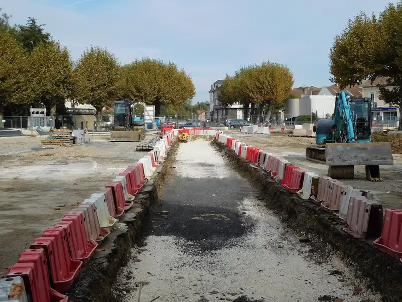 Longue tranchée ouverte entre deux rangées de barrières de chantier sur une place, avec une pelle à l'arrêt à droite
